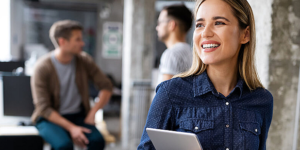 Eine lächelnde Frau hält ein Tablet in der Hand und steht in einem modernen Büro. Im Hintergrund sind zwei Männer zu sehen, die miteinander sprechen. Die Atmosphäre wirkt freundlich und kooperativ.